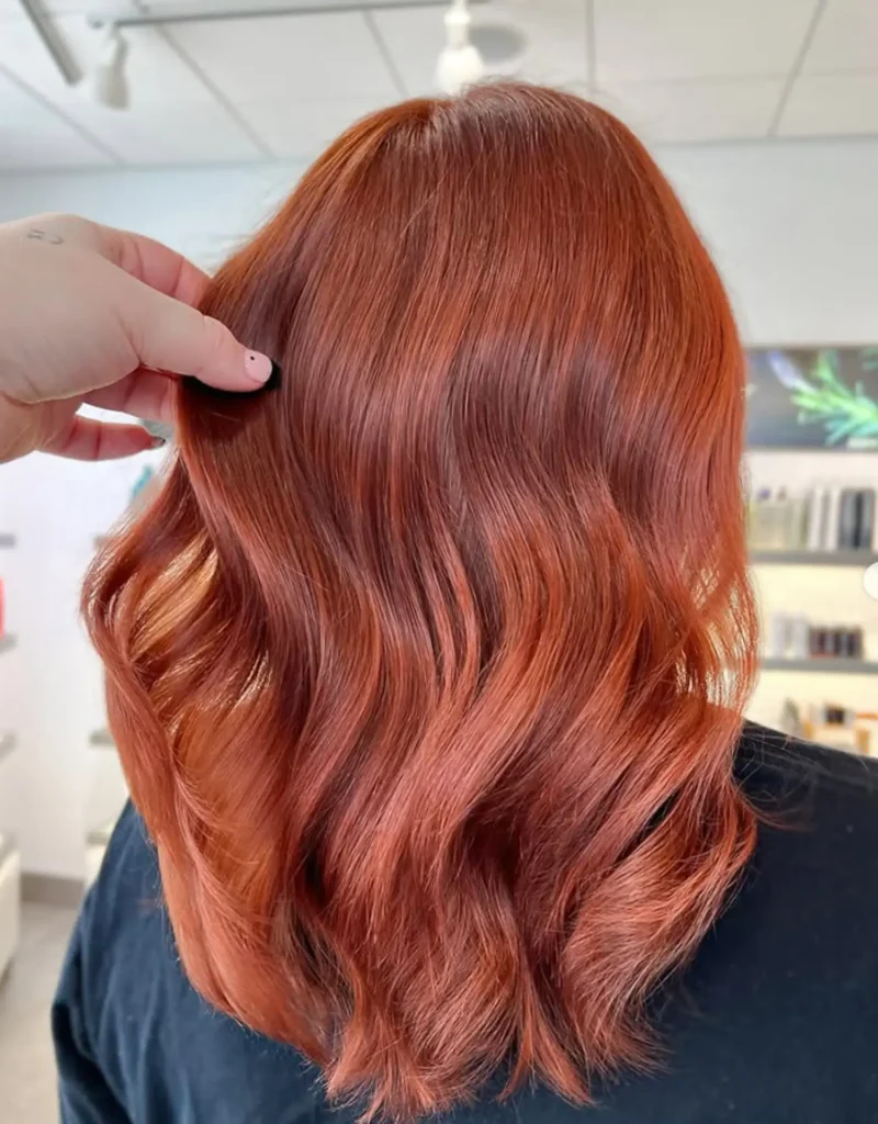 A person with shoulder-length, wavy, copper-red hair is shown from behind in a well-lit salon. A hand is gently lifting a section of their hair. - Mango Salon, VA