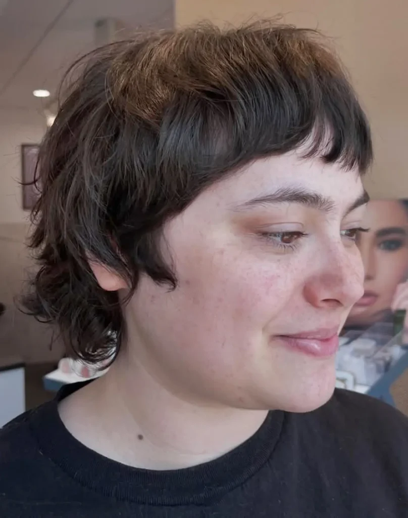 Person with short, wavy brown hair and uneven bangs, wearing a black shirt, looking slightly down and smiling in a well-lit indoor setting. - Mango Salon, VA
