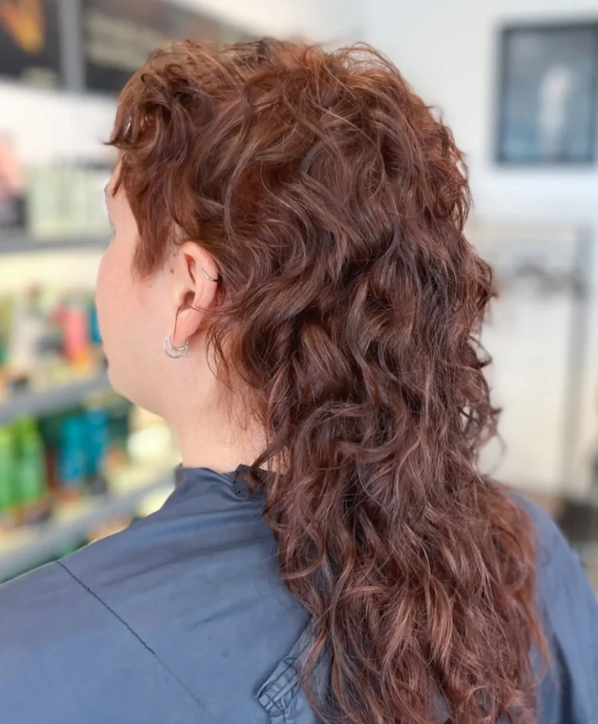 Person with curly, reddish-brown mullet hairstyle sits in a salon chair, seen from behind. Shelves with hair products are blurred in the background. - Mango Salon, VA