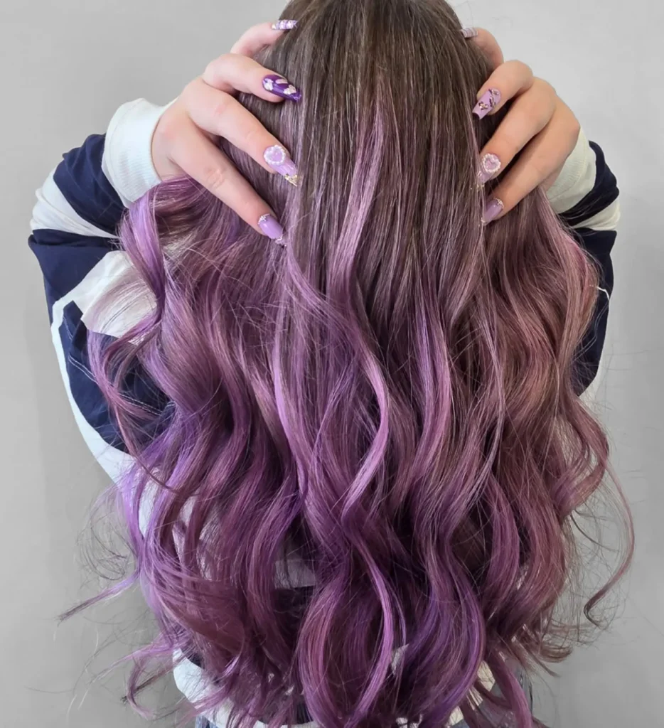 Person with long, wavy, purple ombre hair and floral nail art holds their head with both hands, facing away from the camera against a plain background. - Mango Salon, VA