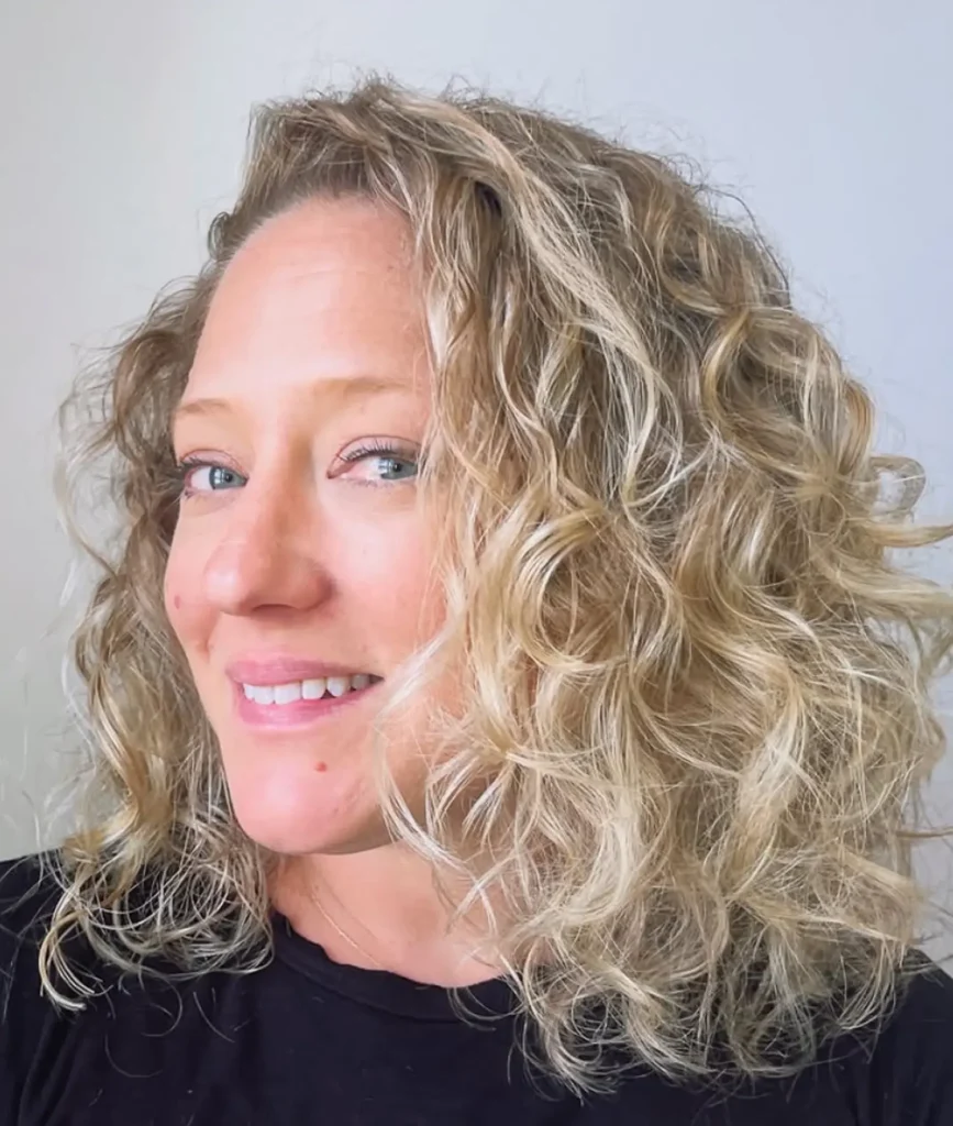 A person with light curly hair and blue eyes smiles slightly, facing the camera with a neutral background. - Mango Salon, VA