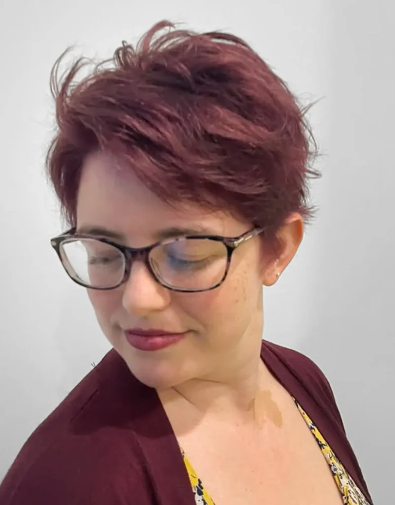 Person with short, wavy, burgundy hair and glasses looks down and to the side, wearing a burgundy cardigan over a yellow patterned top against a plain background. - Mango Salon, VA