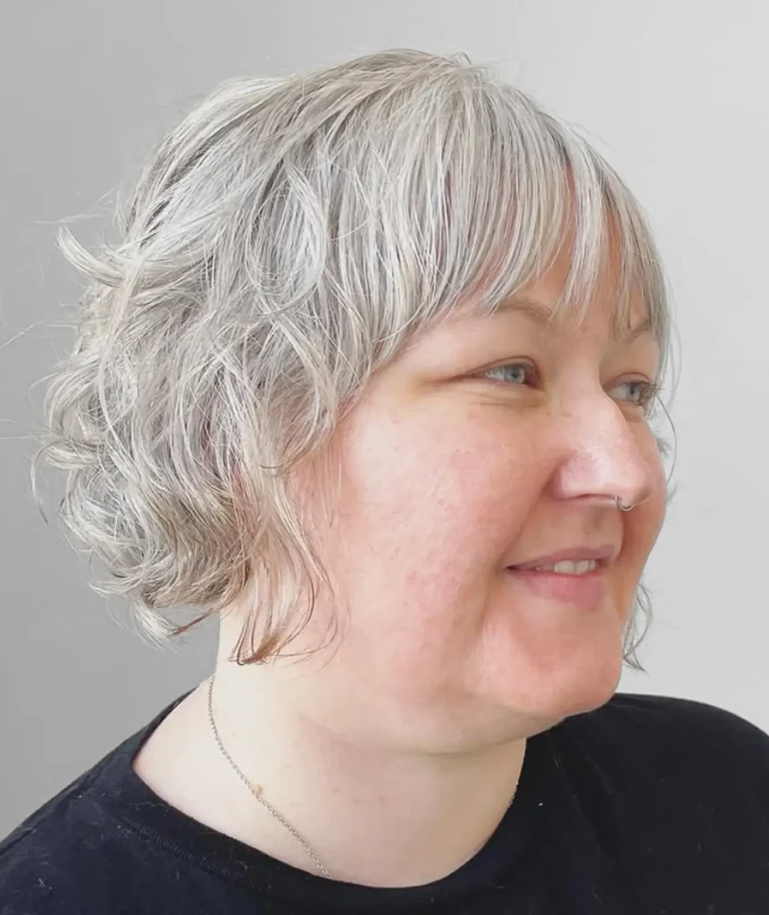 A person with short, wavy gray hair and a nose ring is smiling, wearing a black top, and facing right against a plain light background. - Mango Salon, VA