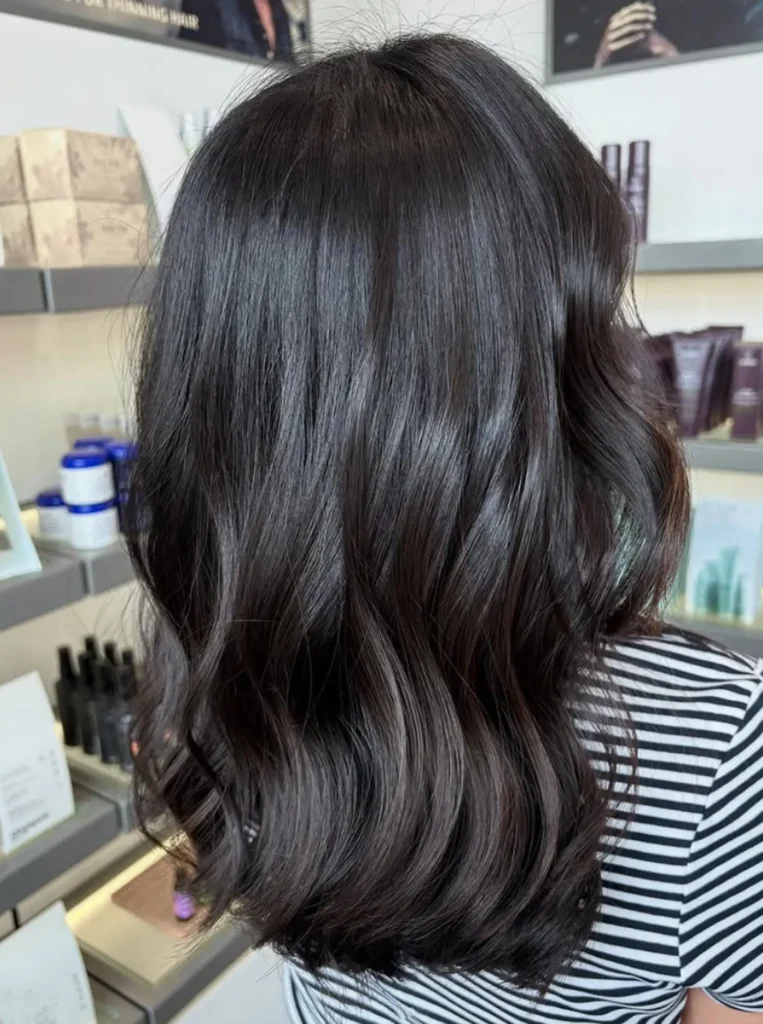 Person with shoulder-length, wavy dark brown hair, photographed from behind in a well-lit store with hair products on shelves. - Mango Salon, VA