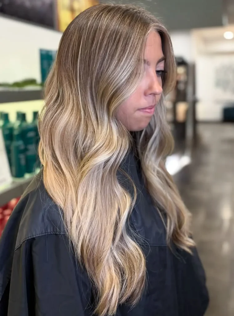 Woman with long, wavy, blonde balayage hair sits in a salon chair, wearing a black protective cape, facing sideways. Shelves with hair products are visible in the background. - Mango Salon, VA