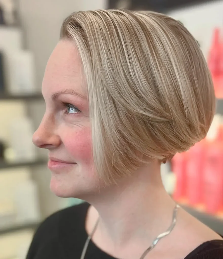 Woman with straight blonde bob haircut, side profile view, wearing a black top and silver necklace, standing indoors with blurred shelves in the background. - Mango Salon, VA