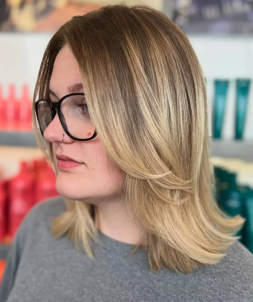 Person with shoulder-length, layered blonde hair and glasses, wearing a grey top, shown in a salon setting with colorful bottles in the background. - Mango Salon, VA