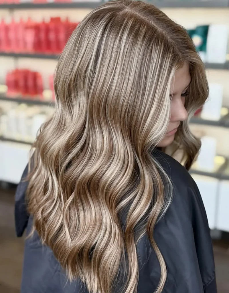 Woman with long, wavy, highlighted blonde hair wearing a black salon cape, background shows shelves with hair products. - Mango Salon, VA