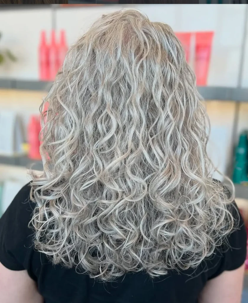 A person with long, curly gray hair is shown from behind, standing indoors in front of shelves with colorful bottles and blurred objects. - Mango Salon, VA