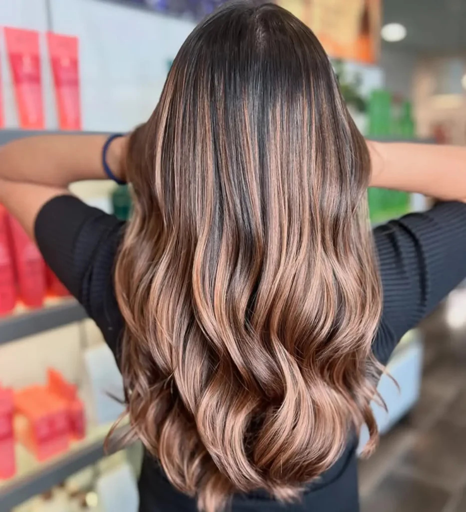 Person with long, wavy, balayage hair in light brown and blonde shades, standing indoors with hands touching head, back facing the camera. - Mango Salon, VA