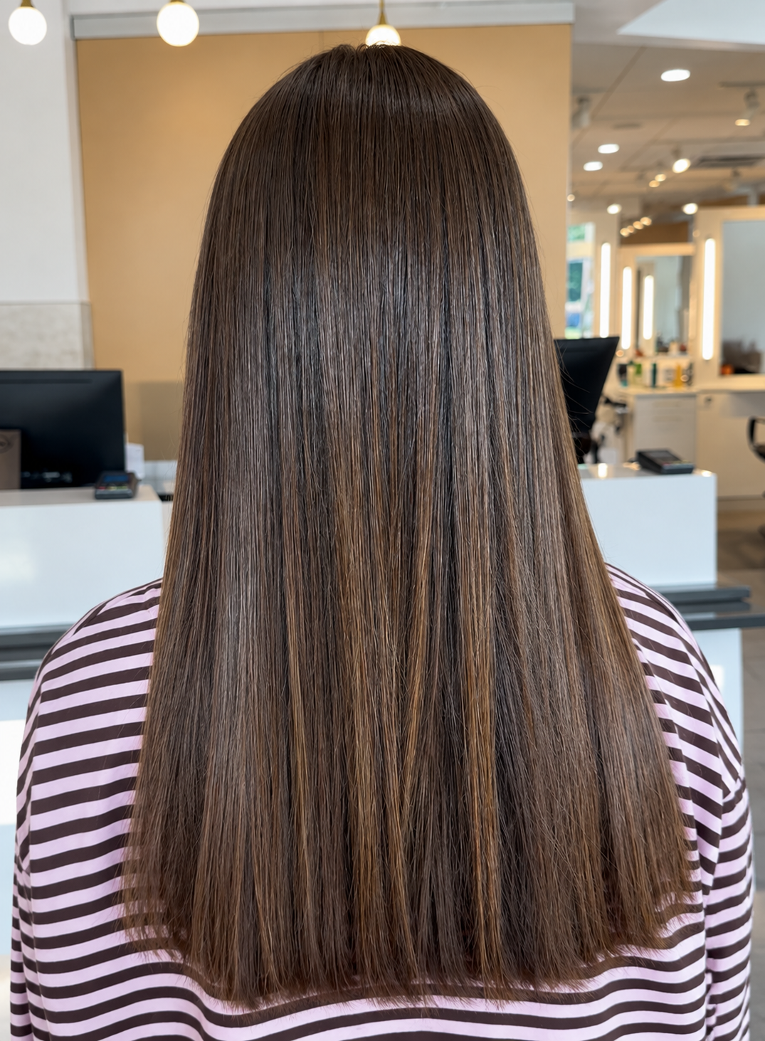 Person with straight, shoulder-length brown and gray hair stands outdoors facing away, showcasing the subtle shine often achieved through restorative treatments, with trees and rooftops in the background. - Mango Salon, VA